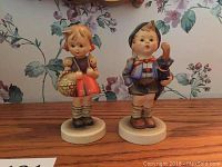 Front view of the pair of Goebel Hummel figurines, girl with basket and boy with umbrella with pig basket.