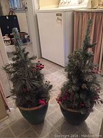 Pair of artificial evergreen Christmas trees with red berry and pine cone decorations in ceramic pots, shown indoors on a tiled floor.
