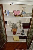 Wooden cabinet shelves displaying signed ceramic vase, colorful mugs, crystal and glass items, brass lion head locks, bar set pieces, and decorative boxes and figurines.