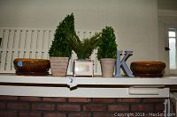 Full view of shelf with two faux green fern plants in ceramic planters, two ceramic bowls, small framed photo, and letter K decorative piece.