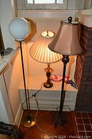 Three lamps: brass table lamp with pleated shade, and two floor lamps with one having a white globe shade and brass base, the other a brown fabric shade with decorative base, arranged near a window and a mantel.