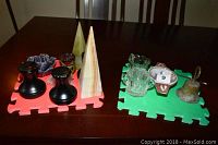 Full view of assortment shown on brown wooden table, two foam pads holding items grouped by color and type, agate obelisks, black decorative items, glass sugar and creamer, bronze bell, rose quartz dragon visible