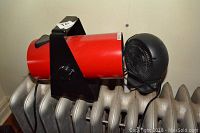 Red Pelonis fan forced heater with black metal stand placed on a radiator, showing front with fan and grille.