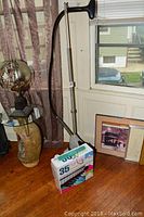 Photo shows the Jiffy clothes steamer model J2 standing upright next to a box with the 35 non-slip clothes hangers in front on a wooden floor by a window.