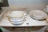 Three Corningware casseroles stacked with lids alongside plain white plates and a clear glass bowl