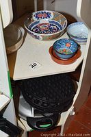 View of decorative bowls including the Kutani bowl and other patterned ceramic bowls placed on shelf, with dark round waffle iron below