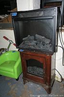 Electric fireplace heater insert placed inside a wood cabinet with glass door and faux logs inside, green plastic chair beside it.