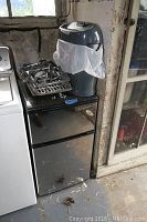 Mini refrigerator with black finish and closed doors alongside adjacent washer. Kitchen appliance grates and a gray Staples paper shredder placed on top.