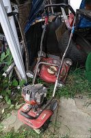 Troy Bilt tiller shown with red body, black handlebars, weed overgrowth nearby, and other yard items surrounding it.
