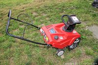 Front-side view of red snow thrower on grass showing handle assembly and left wheel