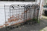 Ten decorative metal garden fence panels with leaf motif stacked and leaned against a wall outdoors.