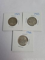 Three Canadian quarters from years 1964, 1965, and 1966 mounted in white coin holders with hand-written year labels.