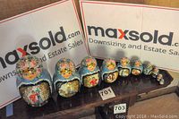 Full view of all ten nested Russian nesting dolls arranged in a line by size on a wooden surface with MaxSold backdrop.