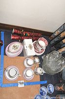 Wide view of assorted tea cups, saucers, decorative plates, small teapot, creamer, sugar bowl, and clear glass bowls grouped on floor.