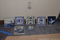 View of seven blue and white decorative plates and two small ceramic pieces arranged on carpeted floor showing varied historical and scenic designs