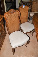 Photo showing two side chairs with hand-carved wood and cane backs, padded seats with light floral upholstery, placed on carpet near other wooden furniture