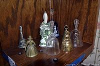 Seven assorted decorative bells arranged on wooden shelf, varying materials and shapes.