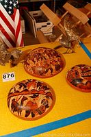 Two metal rooster figurines and five carved decorative wooden bowls with painted scenes, displayed on a yellow surface.
