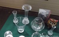 Photo showing assortment of clear glass candle holders, vases, and decorative items with owl figurine on green surface, depicting lot contents.
