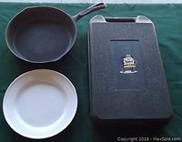 Cast iron skillet, enameled steel plate, and plastic BBQ tool case together on a green surface.