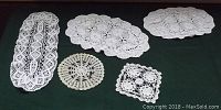 Five assorted white crocheted doilies of various shapes on a dark green surface.