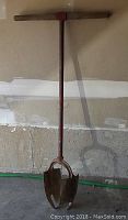 Front view of vintage red metal post hole auger with hardwood T-handle and double blade metal bit for digging holes, standing vertically against a concrete wall.