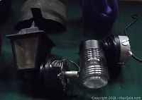 Three outdoor lamps with black metal housing and glass panes, along with a purple watering can and a piece of curved translucent material.