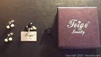 Four pairs of earrings displayed on dark surface with Feige Jewelry box visible; includes white and black pearl pairs and black steel disc pair.