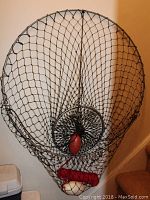 Full view of seining net hanging on wall showing large circular metal hoop and attached black netting, red rope, and buoy floats inside net bottom.