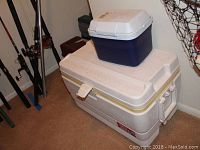 Two coolers stacked; large white Coleman cooler on bottom, smaller blue cooler on top, in a basement by fishing rods and net