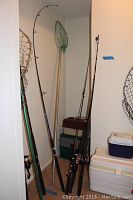 Four fishing rods and one net grouped in corner leaning against the wall next to coolers and storage boxes