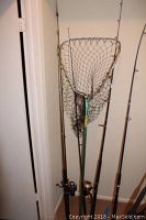 Four fishing rods and one large fishing net leaning in a corner against a white wall on carpeted floor.