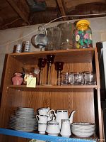 Wooden shelving unit with multiple shelves holding dinnerware sets, glassware, and various vases and candle holders including red stemmed glass candle holders and colored vases.