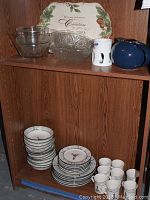 Wooden shelf shelf with stacked plates, bowls, cups and glass serving bowls. Also includes a Christmas-themed serving platter and a blue teapot.