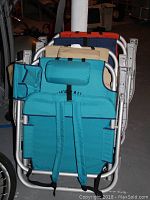 Folded beach chairs in turquoise, beige, and orange colors leaning against a wall in a basement space. Chairs are stacked and show backpack style carrying straps and metal frames.