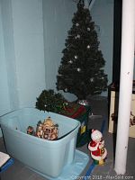 Six-foot artificial Christmas tree with snowflake ornaments and silver stand, green pre-lit tree, plastic bins containing miniature Christmas village pieces and other Christmas decor, plus a ceramic Mrs. Claus figurine wearing red and white.