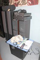 Infinity speaker system components including tower speakers, center channel speaker, subwoofer, Zenith DVD player, and Fisher VCR stacked with cables and cords on a blue cart nearby.