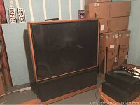 Large 60-inch projection TV unit with wooden trim around screen, black base with speaker covering, situated in basement room with concrete walls and ceiling beams.