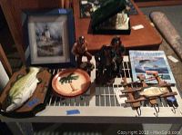 Photo showing Big Mouth Billy Bass mounted fish on wood, wood oar hook with fish and pole, framed lighthouse print, Salt Water Fishing Tactics book, three resin pirate figurines, and two palm tree painted plates arranged on a table.