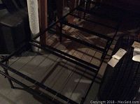 Photo showing the unfolded black metal twin folding cot frame with grid support base set on a floor in a basement.