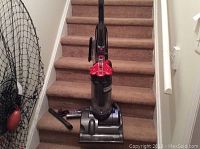 Full view of the Dyson upright vacuum cleaner in red and gray, placed on carpeted stairs with multiple attachments beside it