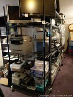 View of large black wire shelving unit loaded with various items including printers and boxes.