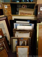 Photo showing a variety of framed and unframed prints stacked against a wall, including large maps and smaller prints with dark and gold frames.