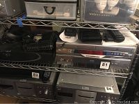 Wide view showing multiple electronic devices on metal shelving including VHS/DVD player and components.