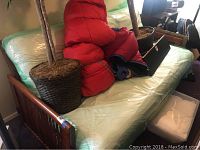 Angle showing wooden framed futon with mattress wrapped in plastic, two artificial potted plants on either end and red sleeping bags on top.