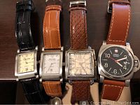 Four square and round analog watches on tabletop: two Prestige, one woven-strap, one Wenger Swiss Military