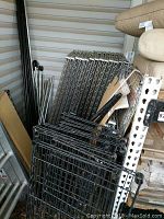 Disassembled silver metal frame sections stacked with black wire shelving panels and tubular frame supports in storage area.