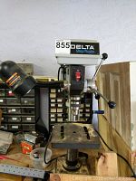 Front view of Delta ShopMaster drill press showing on/off switch, feed lever, chuck, and work table with dust accumulation.