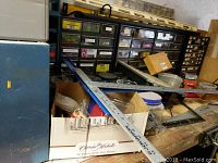 Overview of the wooden work bench covered with T-square measuring rulers, hand tools, cardboard boxes, and plastic jars. Storage drawer units fill the background containing labelled compartments of hardware.