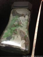 Side view of a glass jar with black lid, filled with green, white, brown, and blue sea glass pieces. A measuring tape shows the jar height to be about 6 inches.